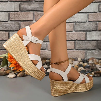 White sandals with gold wedge heels worn by a person against a stone wall background.