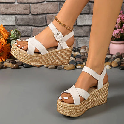 White sandals with woven platforms worn by a person, against a stone wall and pebbles background.