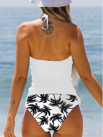 Woman wearing a white tank top and black palm tree patterned high-waisted bikini bottom by the ocean.